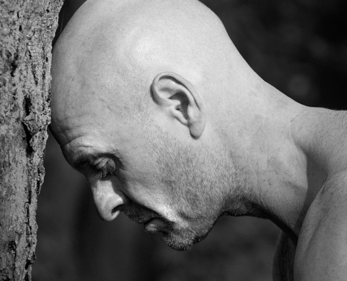 Profilfotografie eines glatzköpfigen Mannes mit freiem Oberkörper, der seinen Kopf gegen einen Baumstamm gelehnt hat.